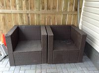 Pair of brown woven patio chairs placed side by side on outdoor stone patio. Chairs are boxy with straight armrests and backs. One chair has arm coming apart.