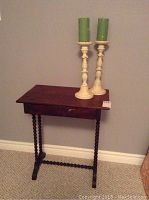 Full view of the wooden table with spindle legs and drawer, showing surface wear and scratches. Two metal candlesticks with green candles are placed on top.