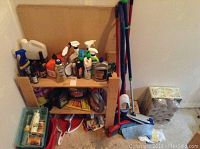 Wooden shelf unit with multiple cleaning supplies and tools including mop, broom, buckets, stacked paper towels.