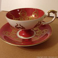 Side view of Aynsley fruit cup on matching saucer with red and gold ornate design and fruit artwork inside cup.