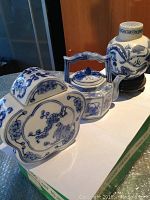 Front and side view of the porcelain tea pot, tea caddy, and vase arranged in a row on a white surface, showing their blue and white decorative designs.