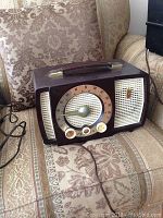 Photo of a vintage Zenith bakelite tube radio placed on a patterned couch. Radio is dark brown with a round dial, three knobs, side speaker grills, and a carrying handle.