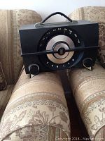 Full front view of the rectangular black bakelite Zenith tube radio showing large circular tuning dial with silver accents, two knobs, and a top fabric handle, placed on patterned couch