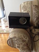 Black vintage RCA Victor tube radio on patterned couch armrest. Shows front grille, tuning dial, RCA Victor logo, and power cord.