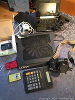 Photo of assorted electronics and camera laid out including Zenit 122K camera, ASUS tablet/netbook, two digital cameras, calculator, and cooling pad