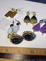 Photo showing multiple pairs of earrings including various stones such as brown, yellow, purple, and opalescent stones, arranged on a white background with ruler for scale.