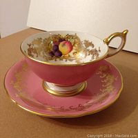 Pink Aynsley cup filled with a multicolored fruit pattern inside, surrounded by intricate gold designs. The cup has a gold-trimmed handle. The matching pink saucer features similar gold decorative filigree around the rim.