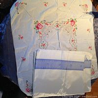 Large and medium sized square linen table covers with colorful floral embroidery, spread over table; one smaller embroidered linen piece with blue hem edge seen on top