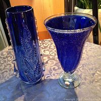 Two blue glass vases on table: one tall cylindrical with leaf pattern, one flared trumpet shape on clear pedestal base