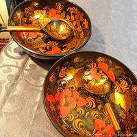 Two wooden bowls decorated with Khokhloma red, black, and gold floral and berry designs, each paired with a similarly decorated wooden spoon inside the bowl.