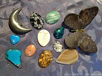 Various polished and carved mineral stones arranged on grey patterned surface including crescent moon, butterfly, hearts, and ovals.