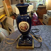 Full view of a blue porcelain table lamp with gold accents on a square gold base, placed on a round table with a patterned tablecloth. Lamp cord visible.