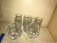 Two clear Waterford Crystal cognac glasses in front with seven clear whiskey glasses behind, all with cut crystal patterns, displayed on a shelf.