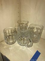 Four Waterford crystal glasses: two classic large rock glasses, one smaller rock glass, and one beer mug with handle, placed on a beige shelf.