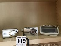 Two silver Olympus Stylus digital cameras and one white Westclox electric alarm clock on a shelf.
