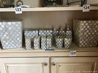 Bathroom accessory set arranged on a shelf, showing various containers, toothbrush holders, dispensers, and cups with a mother of pearl checkerboard pattern.