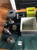 Photo showing three tissue holders in grey, black, and lime green colors, along with multiple silver and black pump dispensers and a small stainless steel cup.