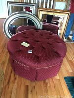 Front view of the large round tufted ottoman in burgundy fabric with button tufting and pleated skirt.