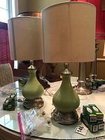 Two light green ceramic table lamps with polished metal bases and beige lampshades on a reflective glass table with assorted light bulbs nearby.