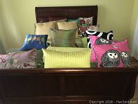 Eleven assorted throw pillows arranged on a wooden bed frame, showing various colors and patterns including owls, florals, and solid shades.