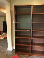 Full view of two brown wood veneer IKEA bookcases side by side, each with six shelves, empty, standing on tiled floor near a wall.