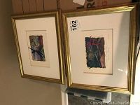 Two framed watercolor paintings resting on a floor, showing abstract colorful artwork and gold frames