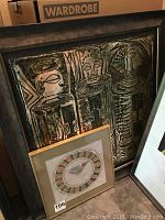 Photo of framed large bas relief metal art depicting three abstract figures with bronze patina and smaller framed Mayan calendar art print placed in front.