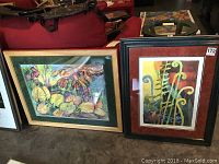 Two framed contemporary wall art pieces placed side by side on floor, colorful botanical themes, one with green mat and gold frame, the other with red mat and black frame.