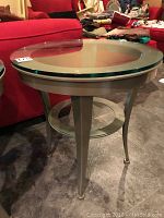 Side view of metal and glass round side table next to red couch showing curved legs and lower circular shelf.