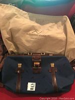 Navy blue weekend bag with brown leather straps and gold-tone hardware placed on a red surface along with protective dust bag with Alessandro Venanzi logo and Made in Italy text.