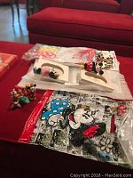 Photo of the red table surface displaying two white picture frames with Mickey and Minnie Mouse figures, small Disney collectible figurines, lapel buttons in original packaging, and folded adult medium Disney T-shirt.