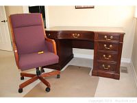Full view of cherry toned executive desk with curved front and multiple drawers beside a rolling office chair with burgundy upholstery and wooden frame.