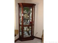 Full view of the wooden corner cabinet positioned in a corner, showing its height, lighting, and contents inside the beveled glass panels.
