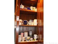 Coffee mugs and ceramic canisters arranged on three wooden shelves inside a cabinet.