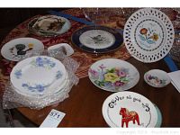 Overview photo showing variety of 15 decorative plates with floral, folk art, bird, and fruit motifs on a round table covered with patterned cloth.