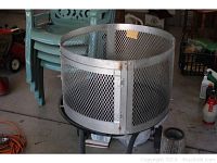 Fire pit metal mesh enclosure shown with four green plastic stackable patio chairs in background.