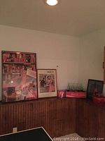 Wide angle photo showing two Coca Cola framed posters, a wooden red Coca Cola crate holding greenish glass soda bottles, a Coca Cola six-pack carton, and a basket of bottle caps arranged on a wooden sideboard.