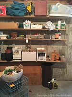 Four shelves filled with gardening supplies including bottles, plant pots, wooden boxes, Sterilite tubs with lids, a black trash bin, and a small animal cage.