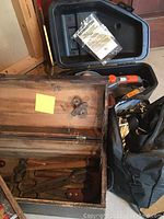 Open view of wooden tool box showing assorted metal hand tools inside, alongside Remington electric chainsaw case with manual on top, and black duffel bag to the right.
