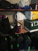 Various black luggage bags and cases piled together with a purple thermos bottle and baseballs visible.