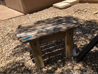 Wooden stool with four legs and natural wood finish placed on gravel ground, showing dimensions sticker.