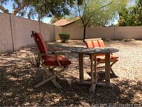 Round wooden outdoor table and two wooden chairs with striped cushions placed outside on gravel ground with natural sunlight. Wood shows faded finish.