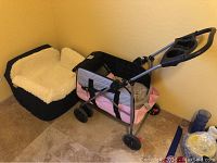 Photo of a black dog car seat with cream fleece inside next to a pink and gray pet stroller with black wheels and handle with cup holders.