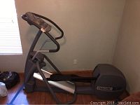 Side view of gray Precor elliptical machine showing overall structure and size in a room with wood floor and taped boundaries.