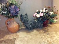Full view of two artificial plants in distinct vases and dog statue on tiled floor against wall.