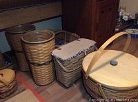 Seven large Longaberger baskets of different shapes including tall cylindrical baskets with metal rims, rectangular basket with fabric-lined lid, and oval basket with wooden lid and handle, placed on wooden floor by furniture.