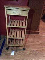 Full view of light wood rolling kitchen cart showing drawer, three slatted shelves, and wheels against a red wall.