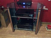 Front view of the TV stand showing black glass top and lower shelves with metal supports and wood veneer side panels.