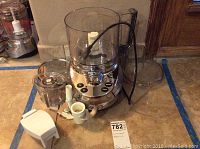 Front view of Cuisinart cast metal food processor with clear bowl and control buttons on base, showing multiple attachments around it on the floor.