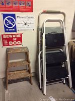 Wooden three-step ladder next to a metal step ladder with black plastic steps and white frame, with wall signs in the background